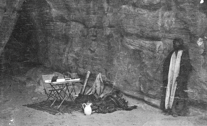 Shakespear’s camping gear and Arab figure by rock face and cave, probably in mountains in vicinity of Wadi Iram/Yatun Aqabah.